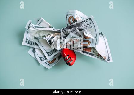 Crumpled hundred dollar bills are secured by heart shaped combination lock on blue background. Secure financial assets, personal savings protection Stock Photo