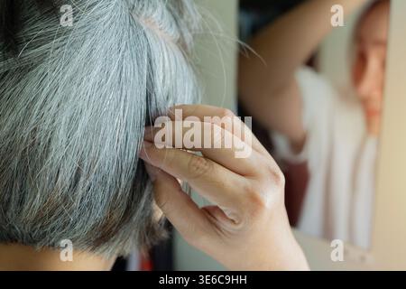 Mature woman with gray hair looking at her reflection in the mirror with thoughtful expression. Close-up, selective focus. Gray hair concept. Stock Photo