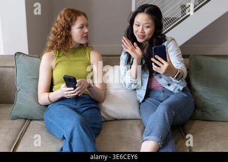 Two women sitting close on beige sofa, holding smartphones, one waving and wearing denim jacket Stock Photo
