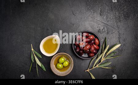 Green and black olives in a bowls and olive oil on dark background top view. Olive oil and variety of green and black olives in bowls with olive leaves all around taken from above. Stock Photo