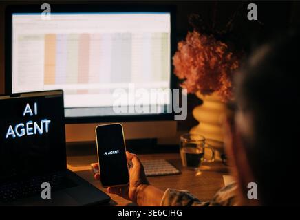 Individual using smartphone with "AI AGENT" text while sitting at a desk with a laptop and computer monitor displaying a spreadsheet and flowers nearb Stock Photo