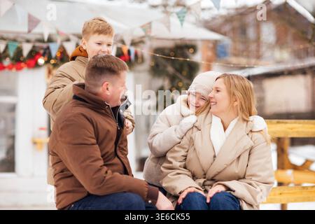 Family of four, including two children, sitting on a wooden bench outdoors in winter, with festive decorations and snow-covered ground surrounding the Stock Photo