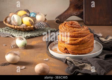 A golden brown cinnamon cruffin sits on a ceramic plate, surrounded by speckled feathers and pastel Easter eggs in a rustic, warm-toned setting with s Stock Photo