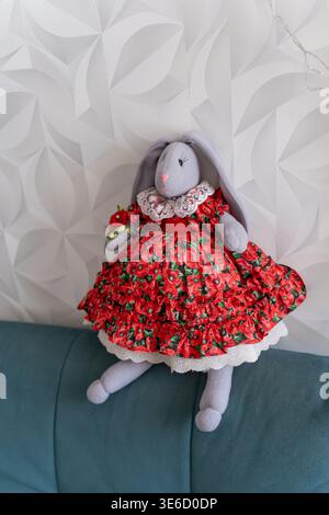 A beautiful, handcrafted plush bunny wearing a vibrant red floral print dress with white lace trim, sitting on a blue surface against a textured white Stock Photo