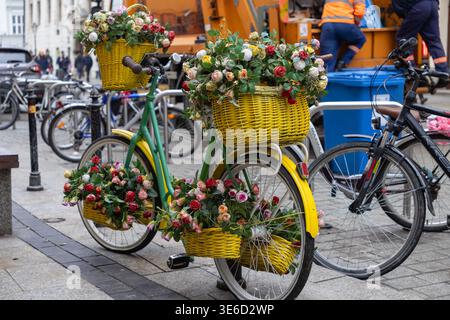 Artistic green bicycle parked on a city sidewalk, displaying yellow wicker baskets filled with vibrant artificial roses, perfect for urban spring deco Stock Photo