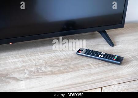 Close-up view of a black television screen with a red standby light and a digital remote control on a wooden cabinet, ready for evening relaxation Stock Photo