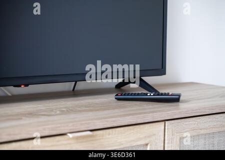 A close-up view of a black television screen with a red indicator light and a remote control on a rustic wooden cabinet, depicting modern media use Stock Photo