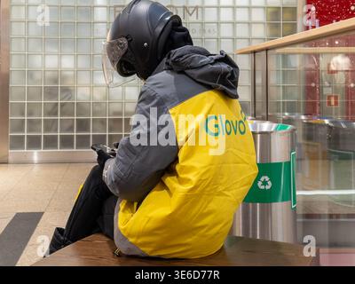 Krakow, Poland - December 2, 2025: Glovo worker in uniform and helmet, sitting, checking smartphone, managing ongoing urban gig economy orders, vital Stock Photo