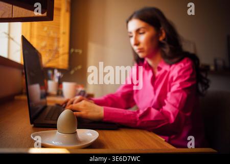 Young woman in pink blouse works on laptop at a wooden desk, featuring a white plate with an egg and warm home office decor in the background Stock Photo
