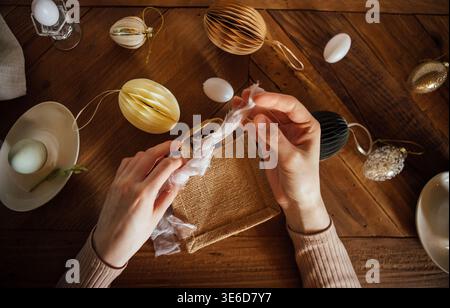 Woman hands carefully wrap a gift with ribbon, surrounded by Easter decorations including colorful ornaments and eggs on a wooden table Stock Photo