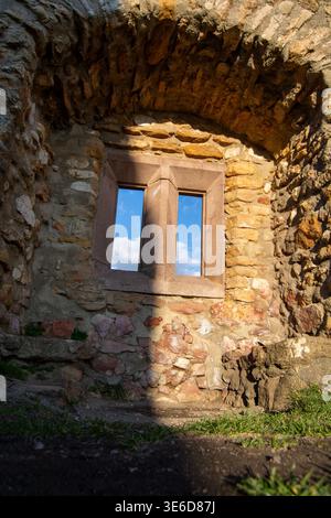 A stone wall with a small window, showing blue sky and clouds outside, sunlight casting a shadow, grassy ground Stock Photo