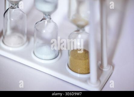 Three hourglasses containing colored sand in white, blue, and yellow, placed on a clean white surface, showcasing a modern and minimalist aesthetic Stock Photo