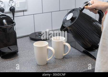 Close-up of a person's hand pouring boiling water from a sleek black electric kettle into two simple white ceramic mugs on a kitchen counter, showing Stock Photo