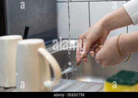 Close-up of a person's hands washing a silver spoon with fresh running water, showcasing routine household tasks and maintaining kitchen hygiene Stock Photo