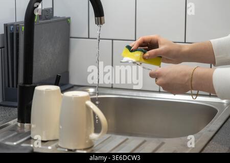 Close-up view of hands scrubbing a fork with a bubbly yellow sponge beneath a stream of water in a modern kitchen, highlighting domestic chores and hy Stock Photo