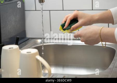 Hands scrubbing a metal spoon with a green sponge under running tap water in a kitchen sink, promoting daily household cleanup Stock Photo