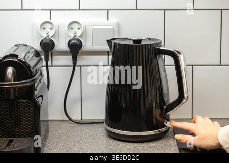 Close-up of a hand activating a modern black electric kettle on a kitchen countertop, with a coffee machine and wall sockets nearby for daily home use Stock Photo