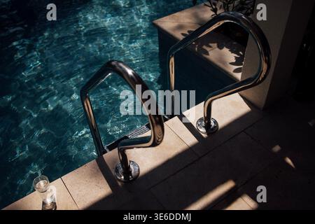 Stainless steel handrails are positioned next to clear blue water, with sunlight creating reflections on the surface and a glass of water resting near Stock Photo