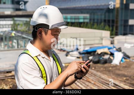 A male engineer in a white helmet uses his smartphone at a construction site under sunlight. He focuses on checking plans and managing work digitally. Stock Photo