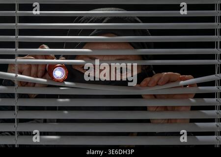 Person with head wrapped in a black scarf and holding a flashlight spying from behind a Venetian blind Stock Photo
