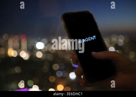 Hand holding smartphone with "AI AGENT" on screen, set against a blurred city skyline featuring colorful lights and urban structures at night Stock Photo