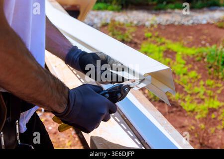 Carpenter uses scissors to cut white piece of vinyl siding while working outdoors on construction project. Stock Photo