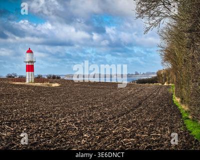 Red-and-white lighthouse stands beside a vast plowed field and shoreline under dramatic cloudy skies, framed by a hedgerow. Rural coastal scene evokin Stock Photo