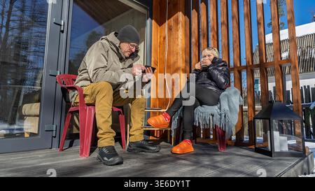 On the terrace of her house, a woman drinks coffee and a man uses a smartphone. Stock Photo