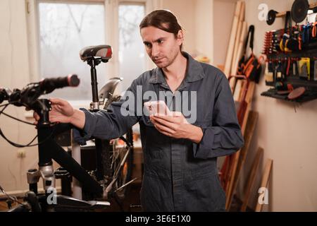 Professional bicycle mechanic in workwear using smartphone while repairing mountain bike in workshop garage. copy space Stock Photo