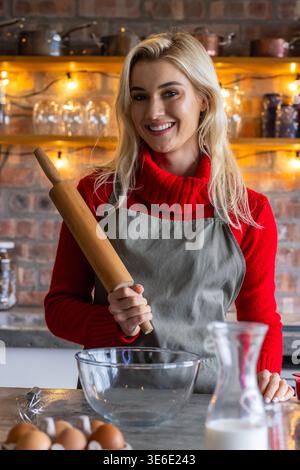 Woman standing at kitchen counter wearing red turtleneck and gray apron, holding rolling pin Stock Photo