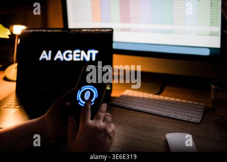 Individual interacts with smartphone featuring biometric security while seated at a desk with multiple computer screens showing data and information Stock Photo