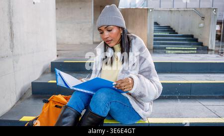 A university student in a cozy sweater and beanie, sitting on campus steps, studies intently with a blue folder and wears headphones around her neck f Stock Photo