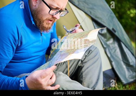 Close up man in glasses closely examines map while holding compass. He sits by his tent, deeply focused on navigation. Lush greenery, suggesting serene outdoor adventure. Stock Photo