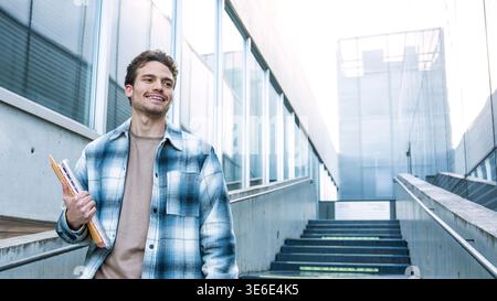 A university student smiles brightly as he holds several textbooks, standing outside on a campus building stairway. The modern architecture surrounds Stock Photo