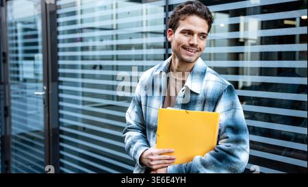 A university student stands outside a modern building, holding a yellow folder and smiling slightly. The glass facade features horizontal stripes, add Stock Photo