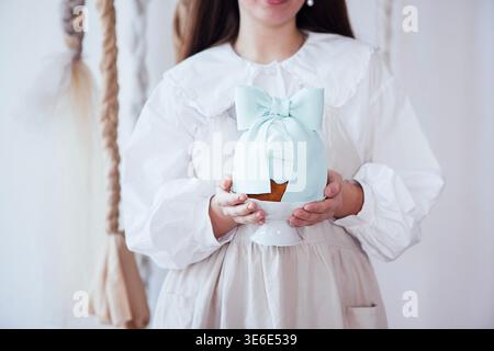 Woman wearing a white dress presents a beautifully decorated cake with a blue ribbon, surrounded by a soft, neutral background with textile elements Stock Photo