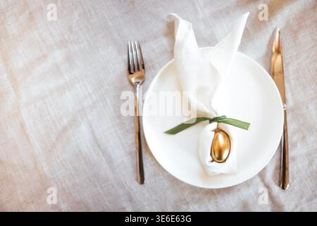 Elegant table arrangement with a white plate displaying a golden egg wrapped in a napkin, flanked by a fork and knife on a linen tablecloth Stock Photo