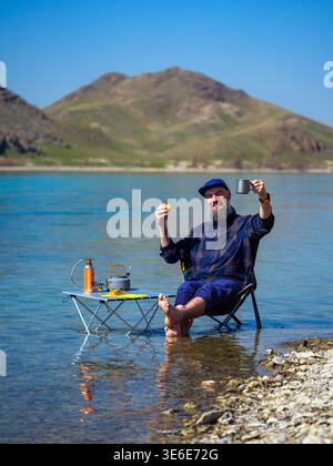Bearded man relaxes in the water near the shore. A cheerful man camps in the river, sitting on a folding chair and drinking coffee Stock Photo