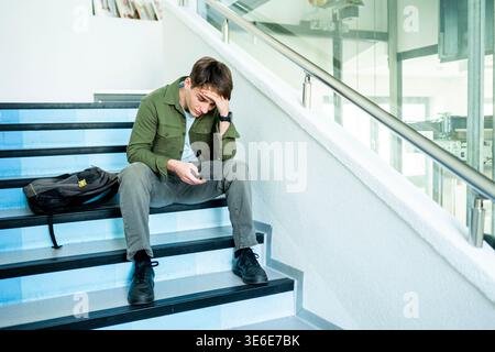 Depressed male student sitting on stairs and looking at phone. Stock Photo