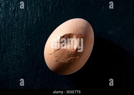 A close up of one brown chicken egg with a dented and cracked shell on a textured dark slate surface. Symbolic image for fragility, beginning, birth, Stock Photo