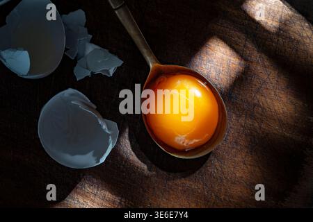 Close up of raw egg yolk in spoon with broken eggshell on rustic wooden surface dramatic light highlighting fresh ingredients and cooking concept. Egg Stock Photo