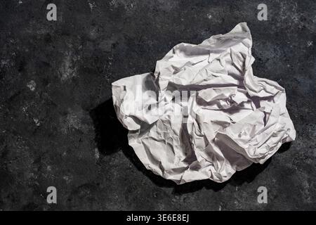 A ball of crumpled white paper on a textured black concrete surface. Symbolic of failed ideas, waste, recycling, or a fresh start in creative business Stock Photo
