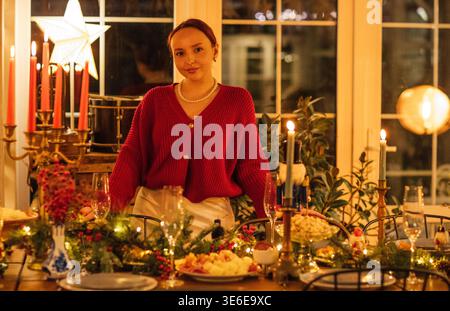 Young woman in a red cardigan poses at a beautifully set dining table with candles and holiday decorations, bright windows creating a warm atmosphere Stock Photo