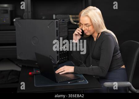 Female IT technician on a call while working on server hardware and laptop in a data center Stock Photo