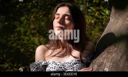 Natural outdoor portrait of a young brunette woman enjoying warm sunlight while leaning against a tree trunk in the forest. Eyes closed, peaceful expr Stock Photo