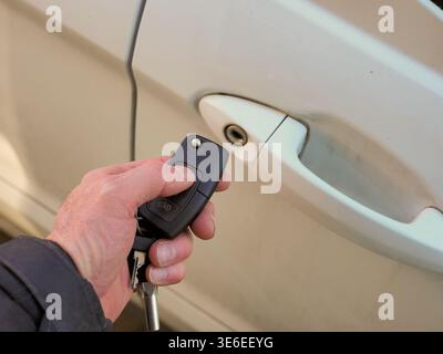 Hand holding modern car key fob with physical key, thumb pressing unlock button to activate remote entry and open white vehicle door, close-up of conv Stock Photo