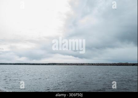 Wide scenic landscape of the Daugava river in Latvia featuring dark ripples on the water surface under a massive dramatic overcast sky with a distant Stock Photo