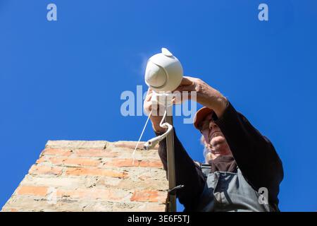 A man in an orange cap installs a white plastic CCTV camera on a brick building under a blue sky. Security system, home technology, outdoor installati Stock Photo