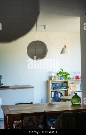 A wooden table, ornate chair, and green glass vase sit under lanterns. A kitchenette leads to a light wood bookcase with books, plant, and a red toy f Stock Photo