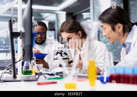 Team of researchers working together, using microscope to evaluate cellular structures in lab. Teamworking colleagues observe medical tissue through magnification tool at laboratory bench workstation Stock Photo